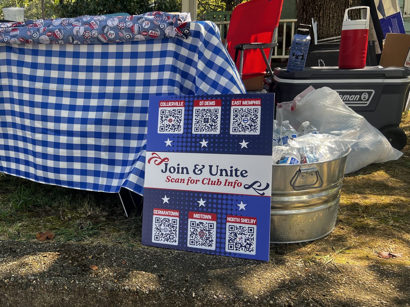 A "Join and Unite — scan for club info" board with QR codes for six Shelby County Democratic clubs, propped beside a tub of iced water bottles.