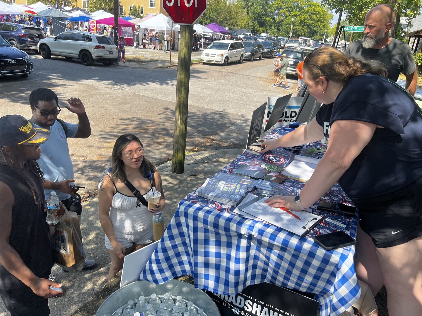 A club volunteer leans across the table in conversation with three festival-goers, the festival's white tents and crowd filling the street behind them.