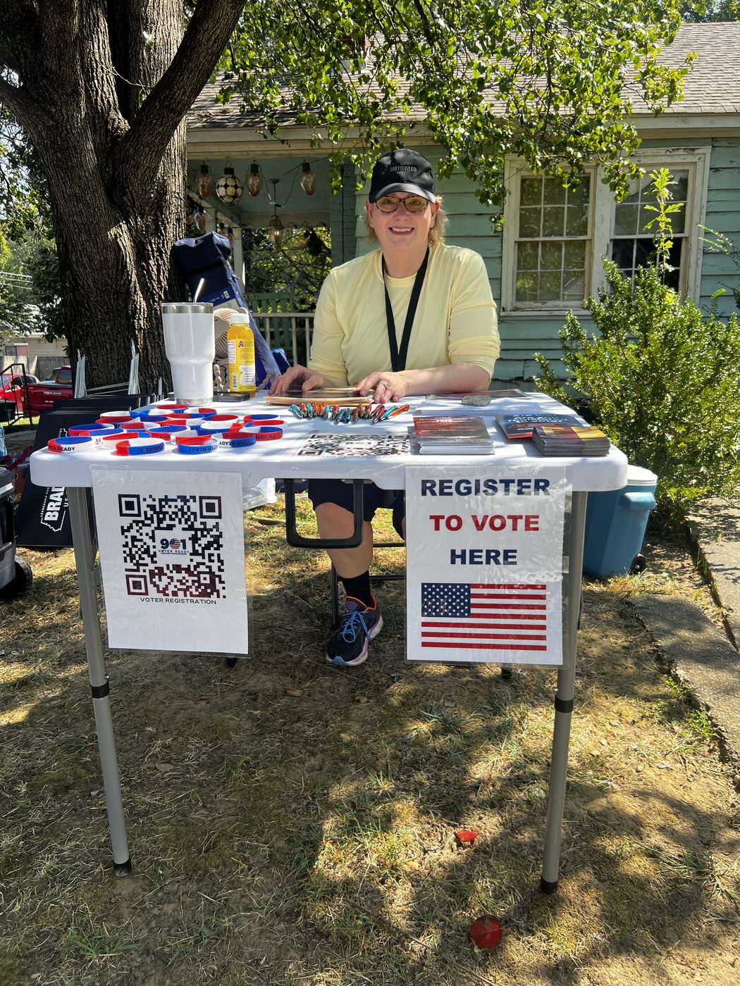 A club volunteer in a cap and lanyard sits smiling behind a folding table marked "Register to vote here", spread with wristbands, literature and a voter-registration QR code.