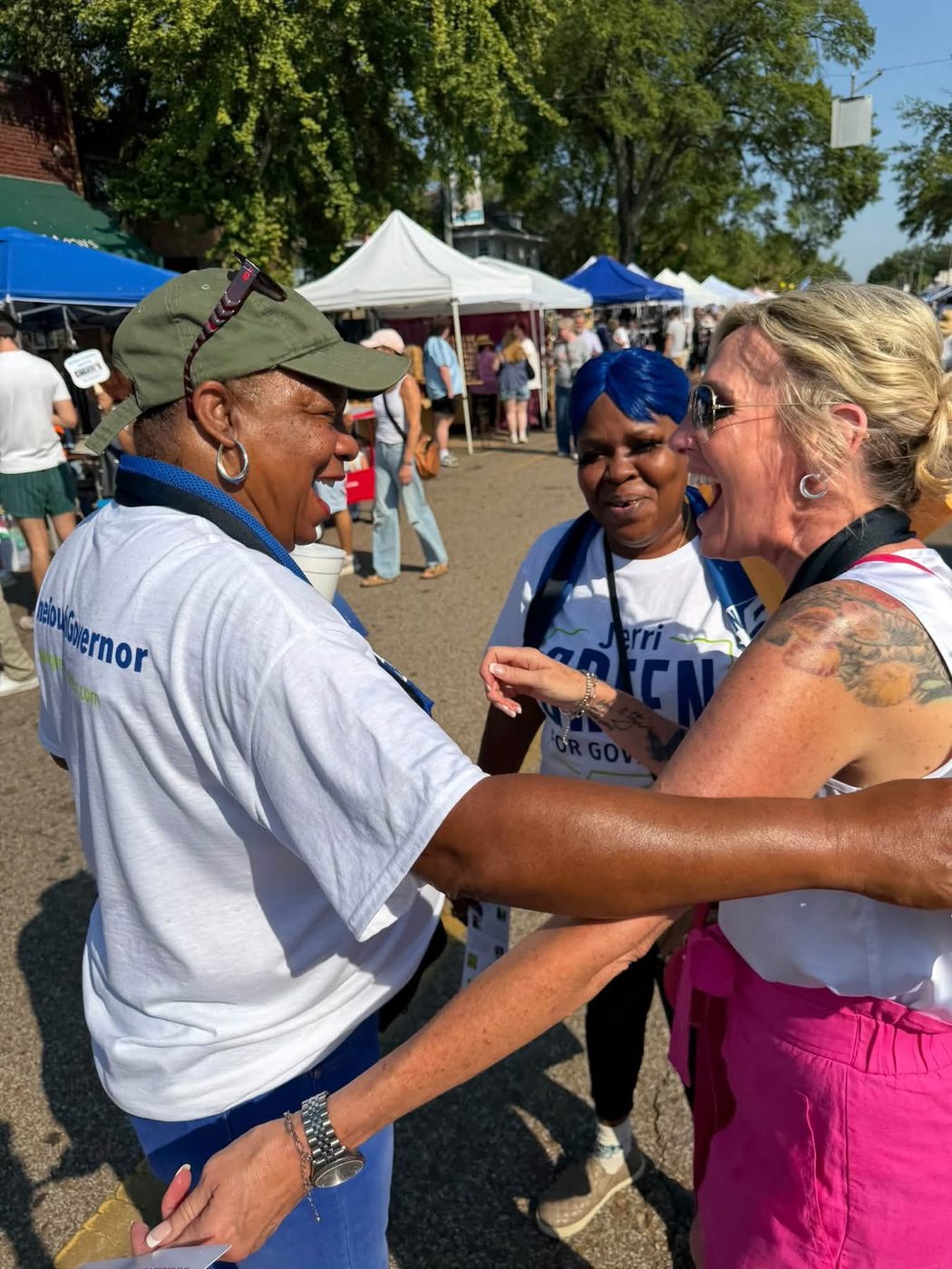 Three women greet each other with laughter in the middle of the festival street, two of them in campaign T-shirts, with tents and crowds behind.