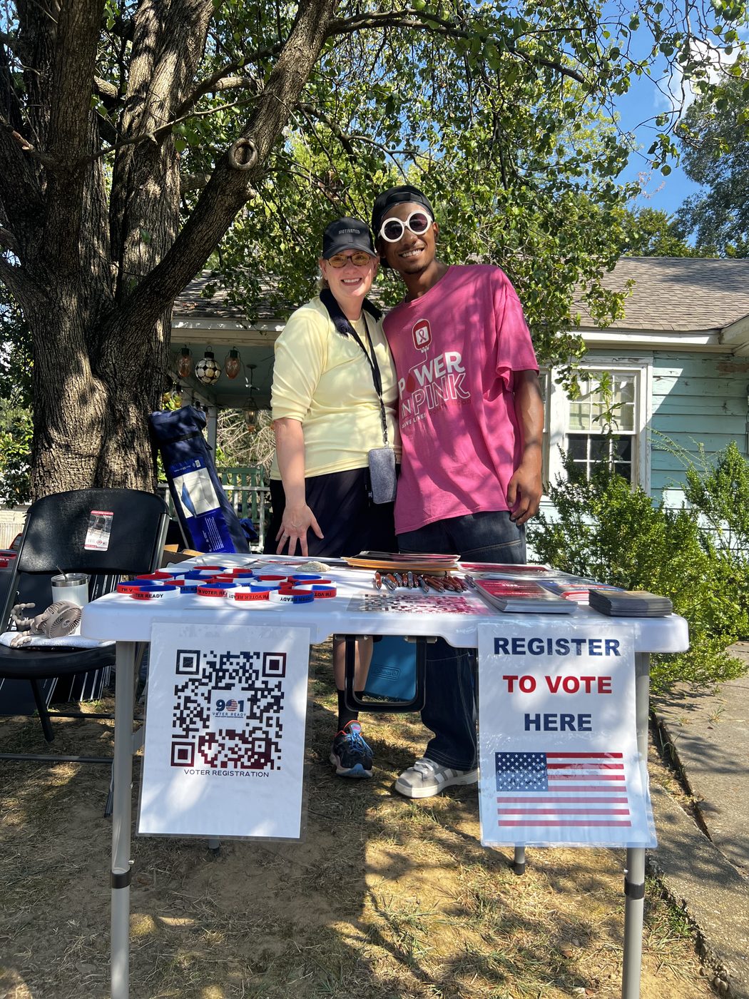 A club volunteer and a young man in a bright pink T-shirt stand arm in arm behind the voter-registration table, both smiling at the camera.