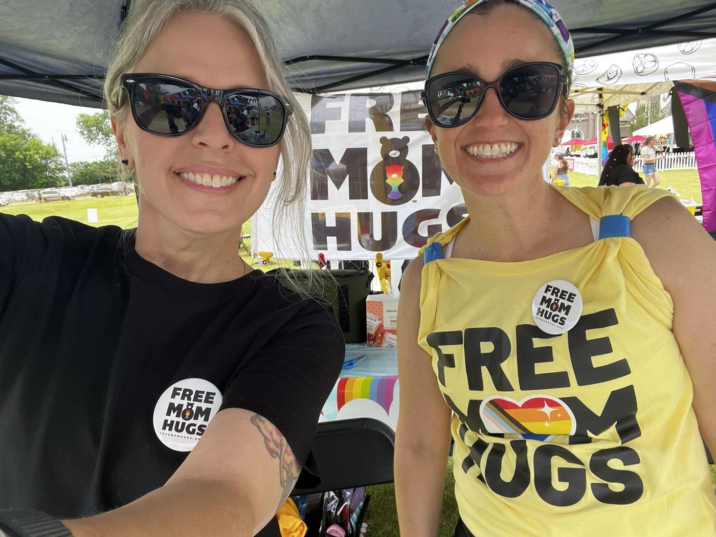 Two volunteers in Free Mom Hugs shirts smiling at the club's tent at a community Pride event.